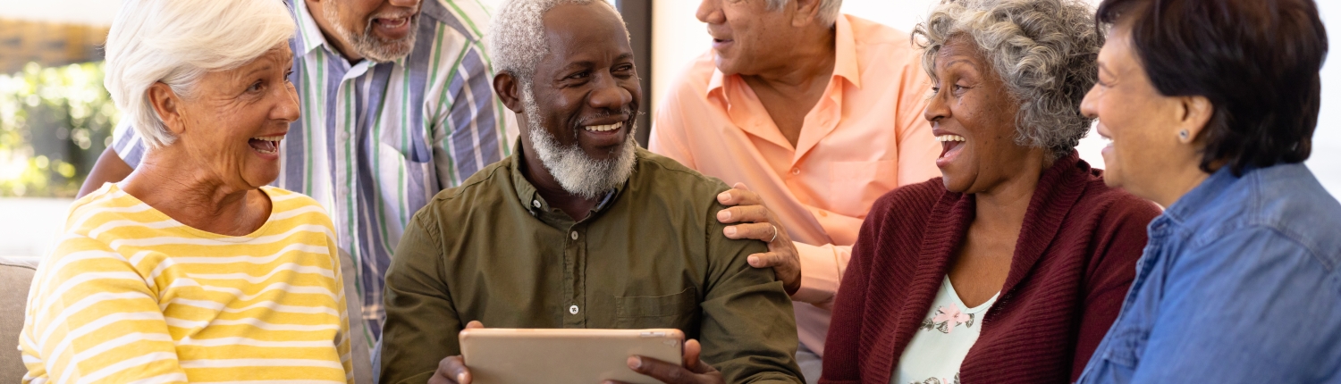 Multiracial senior man holding digital tablet looking at laughing friends while sitting on sofa. Nursing home, wireless technology, unaltered, togetherness, support, assisted living, retirement.